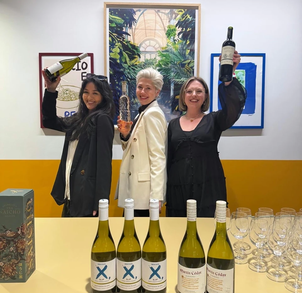 Three women holding wine bottles behind a tasting table with wine glasses and bottles