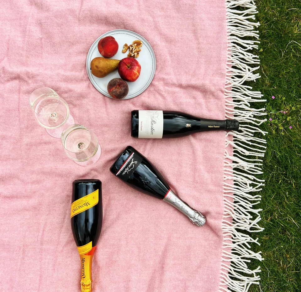 Three Prosecco bottles, two wine glasses and fruit on a pink blanket on grass