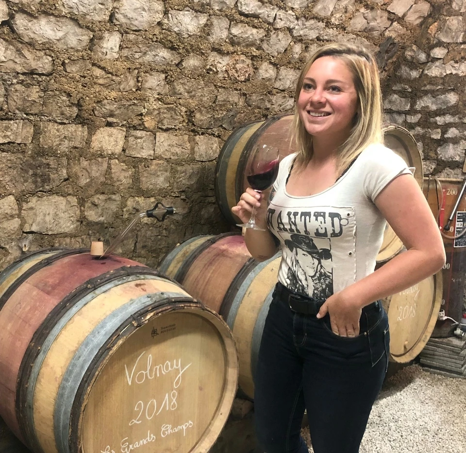 Woman holding a glass of red wine beside oak barrels in a stone wine cellar