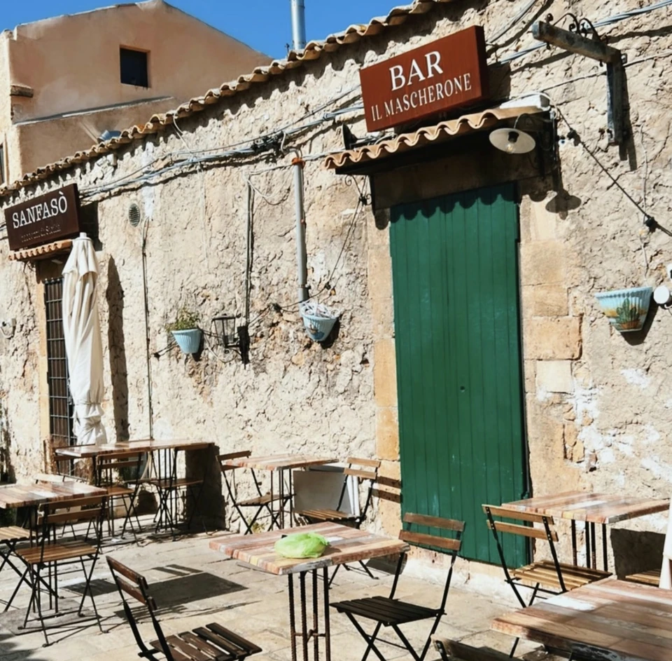 Sunny Sicilian courtyard with outdoor tables and a bar sign reading Il Mascherone
