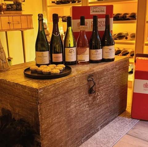 Six wine bottles and savoury tarts displayed on a wooden chest beside illuminated wine shelves