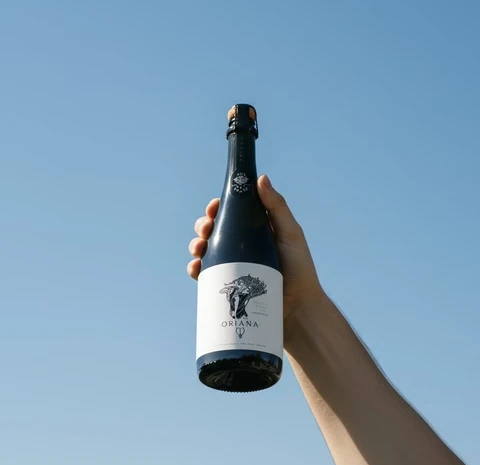 A hand holds an Oriana sparkling wine bottle against a clear blue sky