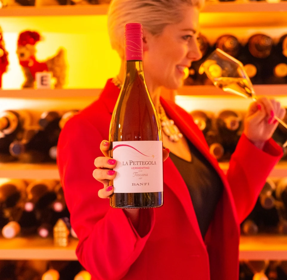 Woman in a red jacket holding a bottle of La Pettegola wine and smelling a wine glass