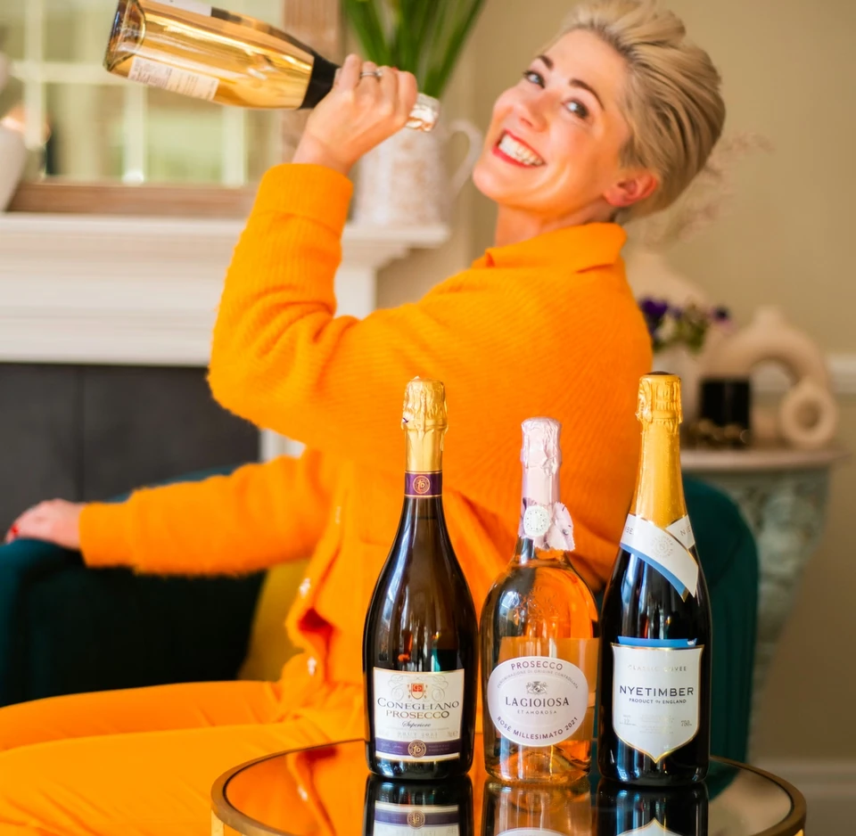 Woman in orange holding a bottle beside three sparkling wine bottles, including Nyetimber