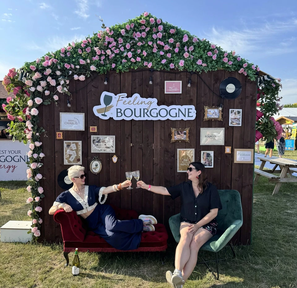 Two women toast with wine glasses beneath a flower-covered Feeling Bourgogne display