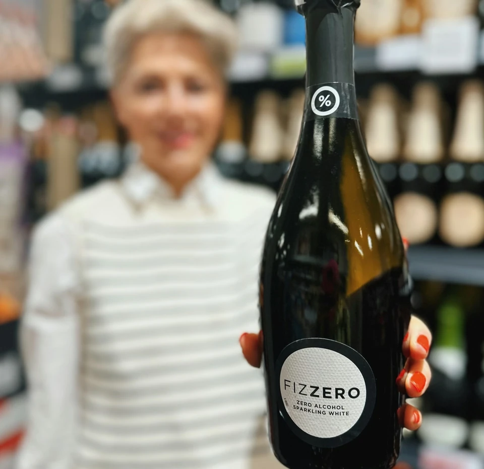Fizzero zero-alcohol sparkling white bottle held in front of blurred wine shelves