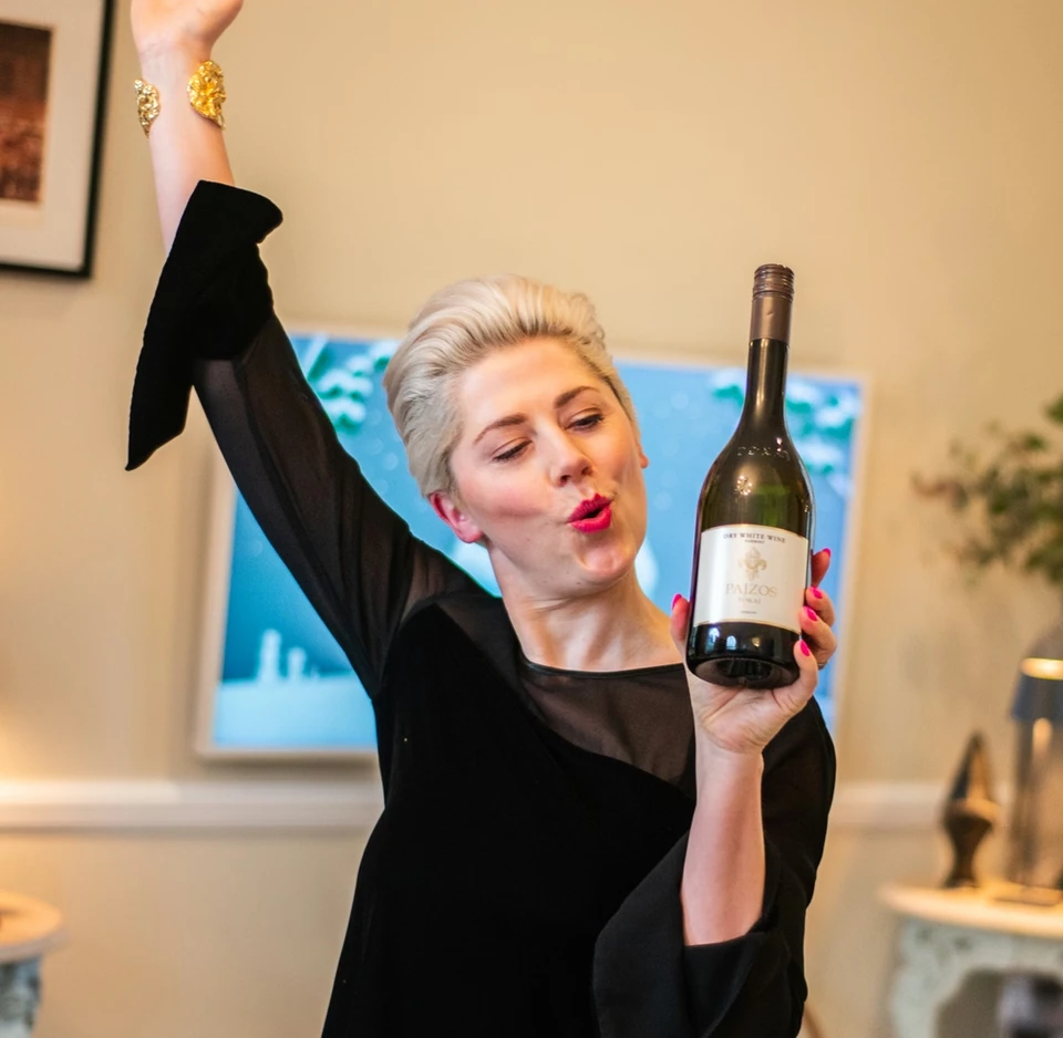 Woman raising one hand while holding a bottle of Paizos dry white wine