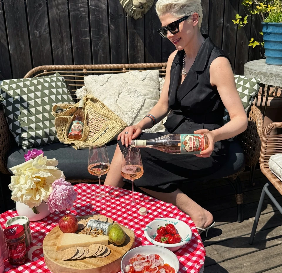 Woman pouring Les Dauphins rosé beside a picnic table with wine glasses, cheese and fruit