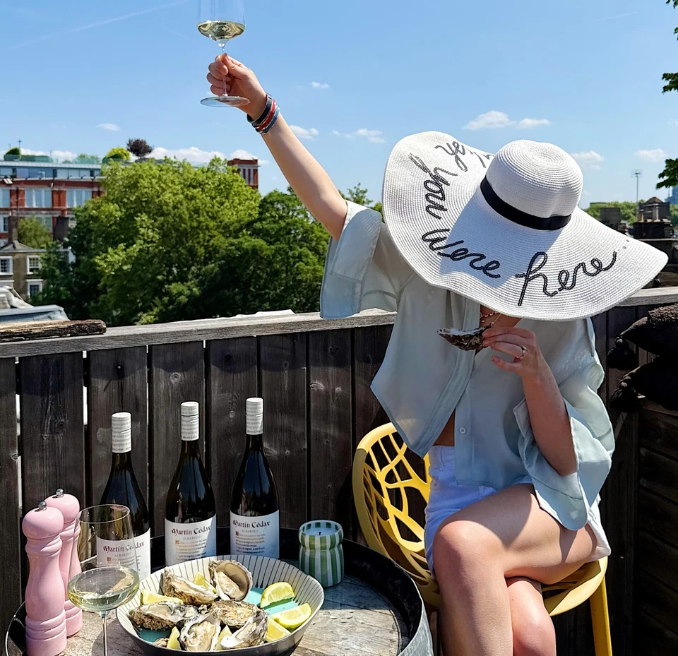 Person in a wide-brimmed hat raising a glass of white wine beside oysters on a rooftop terrace