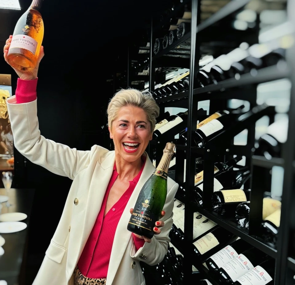 Elizabeth holding two Cava bottles beside a wine rack