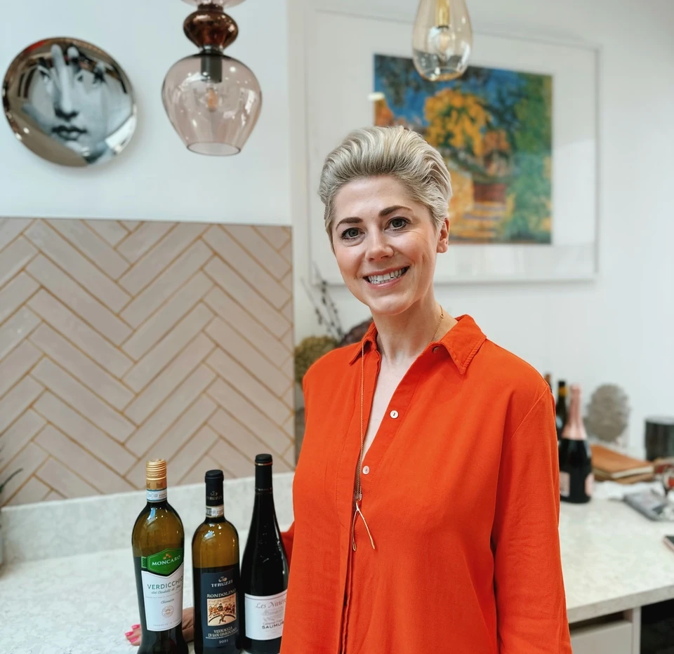 Woman in an orange shirt standing beside three wine bottles in a modern kitchen