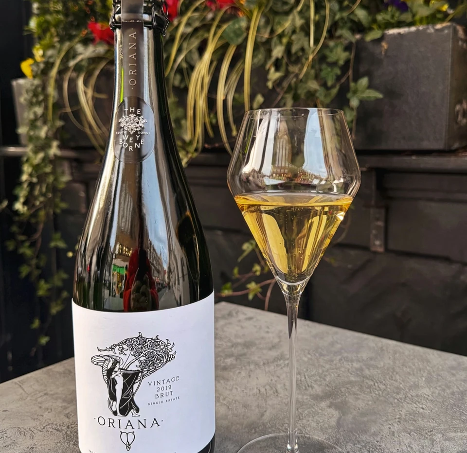 Oriana 2019 Brut bottle beside a glass of sparkling wine on a flower-filled terrace