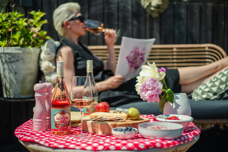 Person drinking rosé beside a bottle of Les Dauphins wine and a cheese board
