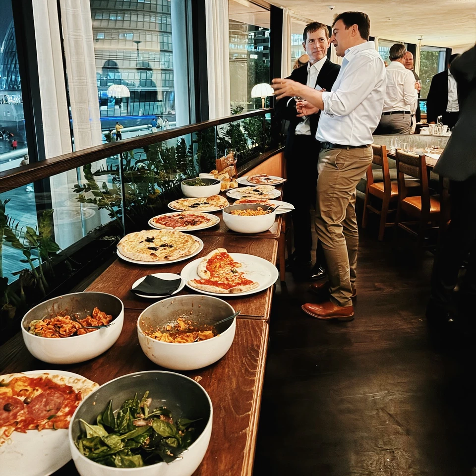 Guests gather beside a buffet of pizzas and pasta in a modern restaurant