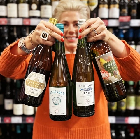Woman in an orange jumper holds a bottle labelled Picpoul de Pinet among other wines