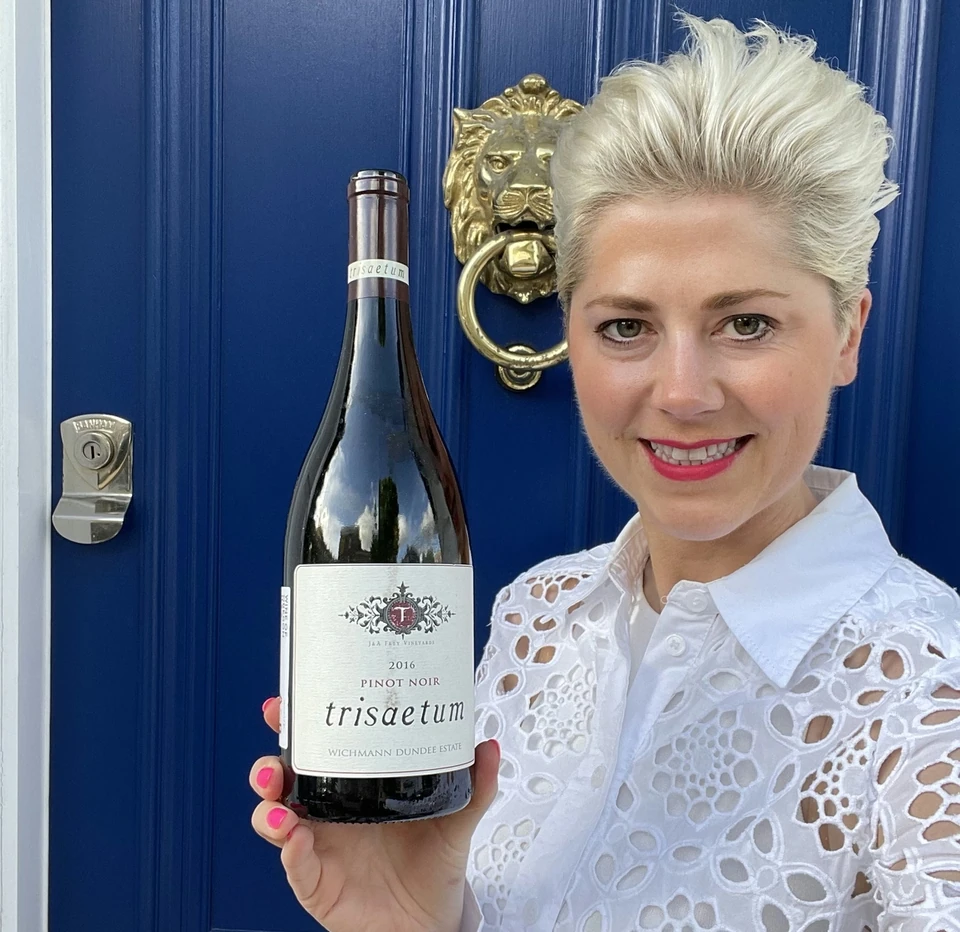 Woman in a white blouse holds a bottle of Trisaetum 2016 Pinot Noir outside a blue door