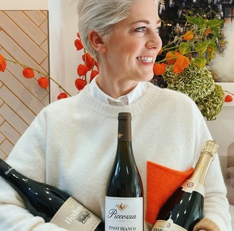Woman in a white jumper holding three wine bottles, including a Pinot Bianco, in a flower-filled kitchen