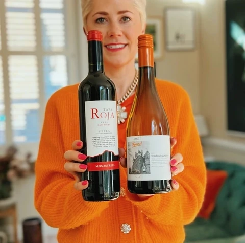 Woman in an orange cardigan holding Tapa Roja and M&S Weißburgunder wine bottles