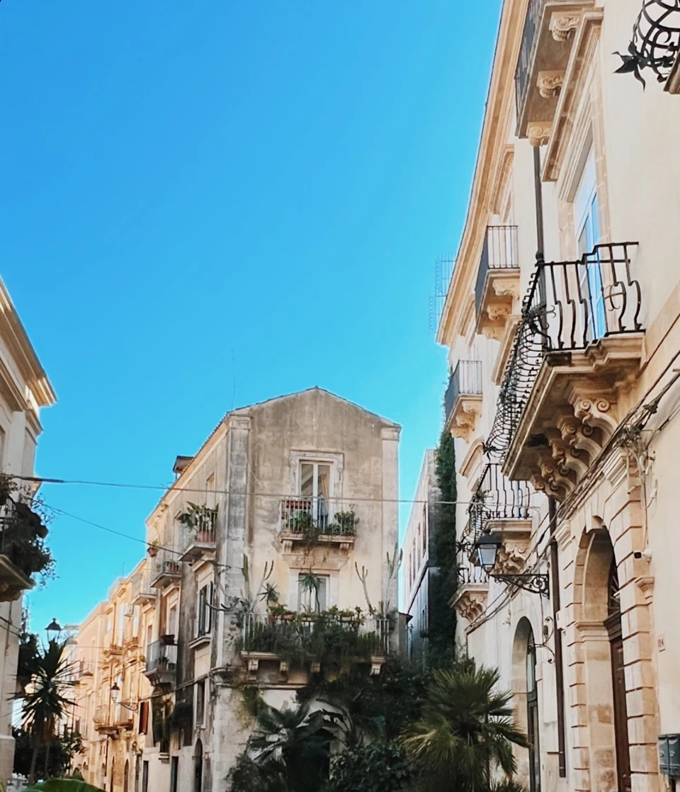 Historic Sicilian street with ornate balconies and Mediterranean plants beneath a clear blue sky