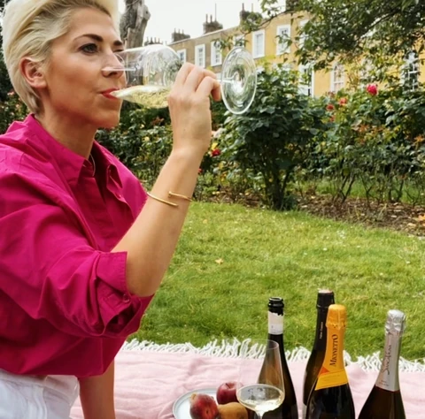Woman tasting sparkling wine beside bottles on a picnic blanket in a garden