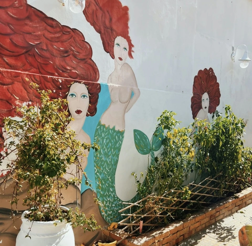 Colourful mural of red-haired mermaids on a white exterior wall with plants and ceramic pots