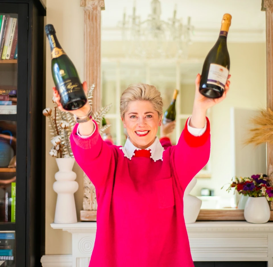 Elizabeth raising two sparkling wine bottles in a sitting room