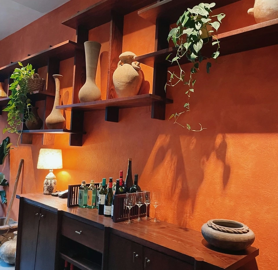 Wine bottles and glasses arranged on a sideboard beneath wooden shelves in a warm terracotta room