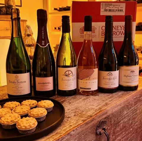 Six English wines and mince pies arranged on a wooden table in front of illuminated wine shelves