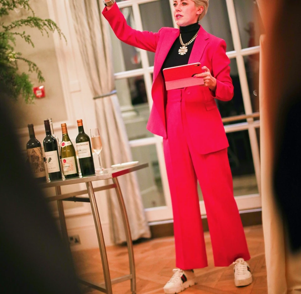 Wine tasting host in a bright pink suit presenting beside bottles of Champagne