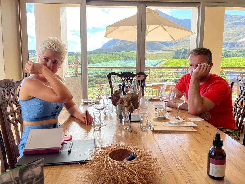 Guests enjoy wine and food at a table overlooking sunlit vineyard hills