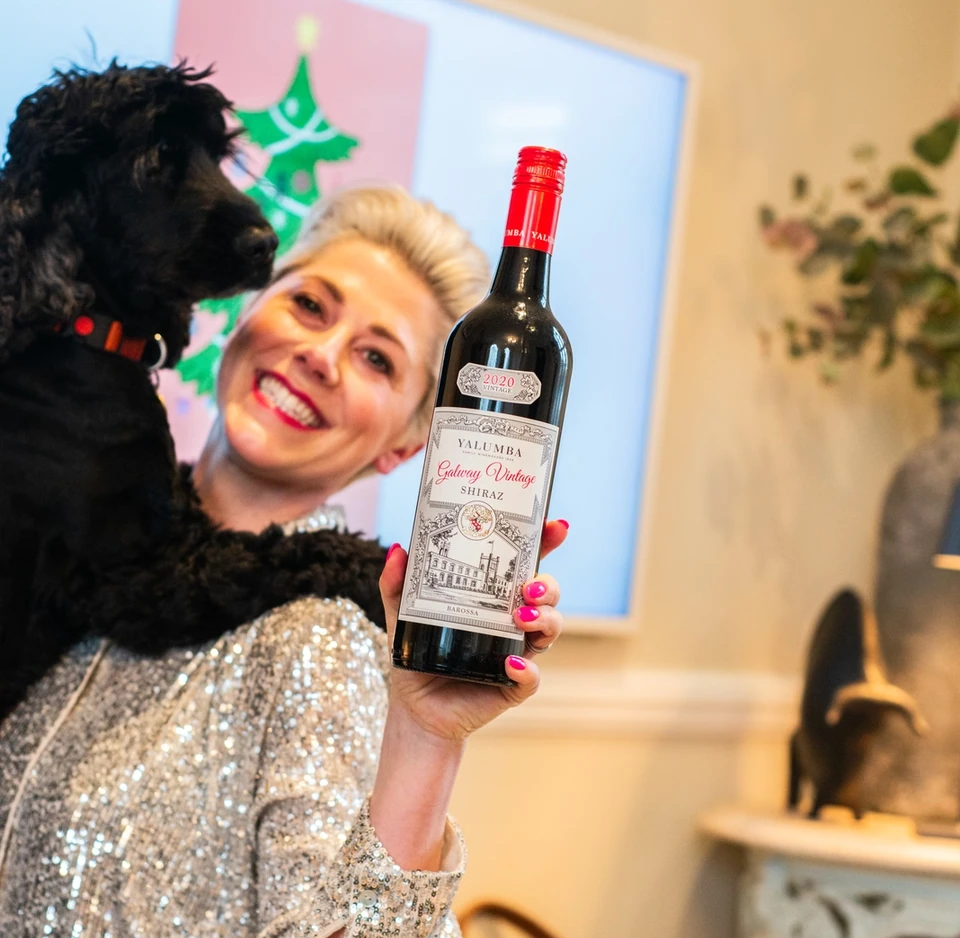 Person in a sequinned top holds a bottle of Yalumba Galway Vintage Shiraz beside a black dog