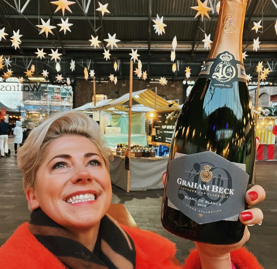 Smiling woman in a red coat holding a Graham Beck sparkling wine bottle beneath hanging stars